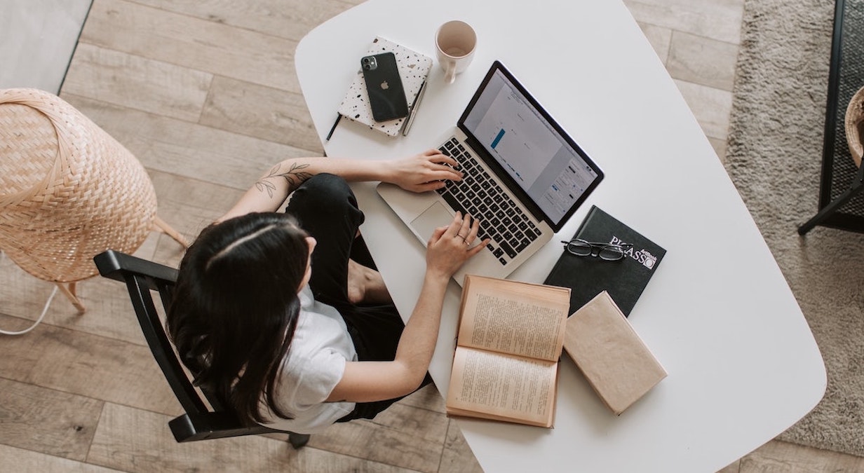 Woman seen from overhead as she looks at a laptop with a book and multiple notebooks beside her.