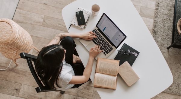 Woman seen from overhead as she looks at a laptop with a book and multiple notebooks beside her.