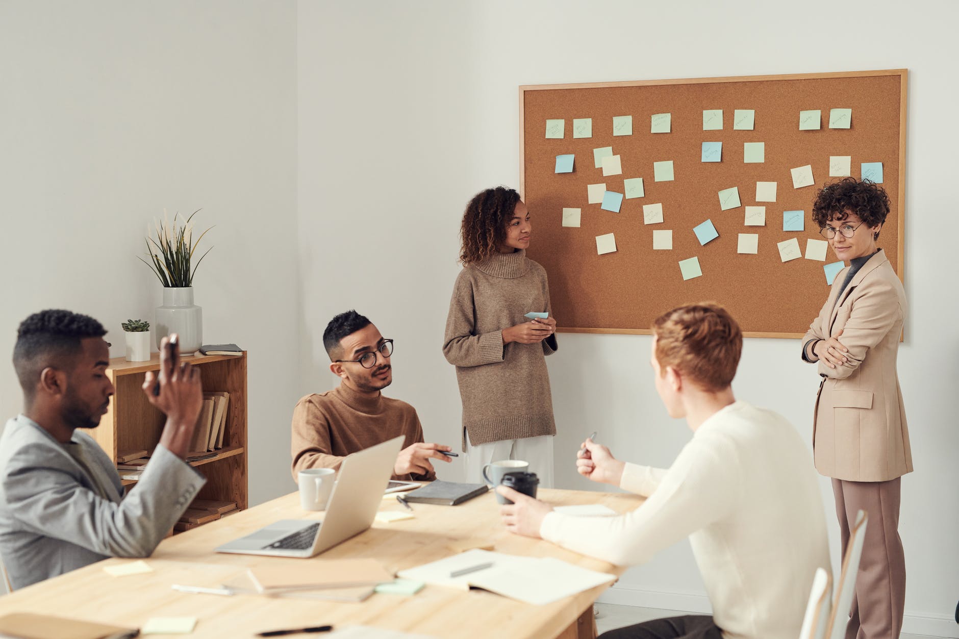 A group of people having a conversation in a conference room. Two people are standing by a bulletin board that has an assortment of post-its on it, and three other people are sitting at the table with a laptop, papers, and coffee.