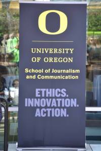Dark stand-up banner has a yellow O and the words "University of Oregon. School of Journalism and Communication. Ethics. Innovation. Action"