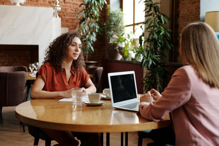 Two women having a conversation while sitting at a table. One has her laptop out while the other listens
